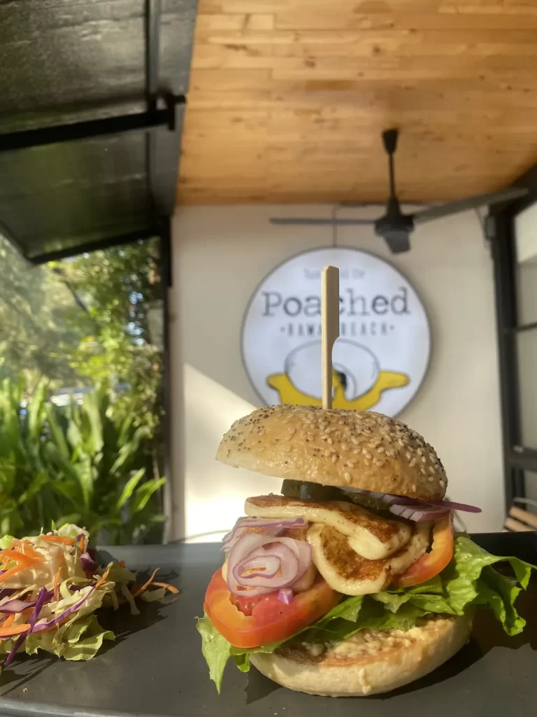 The image is focused on a halloumi bagel topped with lettuce, tomato, and onion with a side salad. In the background is a ceiling fan and the Poached Breakfast Cafe sign. One of the best backpacking activities in Rawai is right here at Poached Breakfast Cafe Rawai, Phuket, Thailand